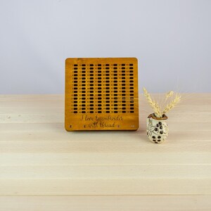 May include: A wooden embroidery hoop with a grid pattern and the text "I love to embroider with thread". The hoop is on a light wood table with a small white vase with dried wheat.