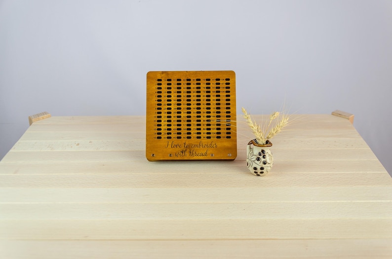 May include: A wooden embroidery hoop with a grid pattern and the text "I love to embroider with thread". The hoop is on a light wood table with a small white vase with dried wheat.