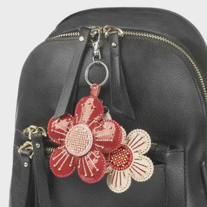 Puede incluir: Una mochila negra con cremallera plateada y un llavero con forma de flor. El llavero tiene dos colgantes de flores, uno rojo y otro crema, con centros de cuentas y detalles de pétalos. El llavero está unido a un anillo plateado.