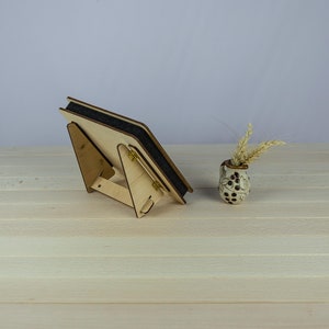 May include: A wooden picture frame with a black backing and a stand. The frame is on a light wood surface with a small vase with dried flowers.