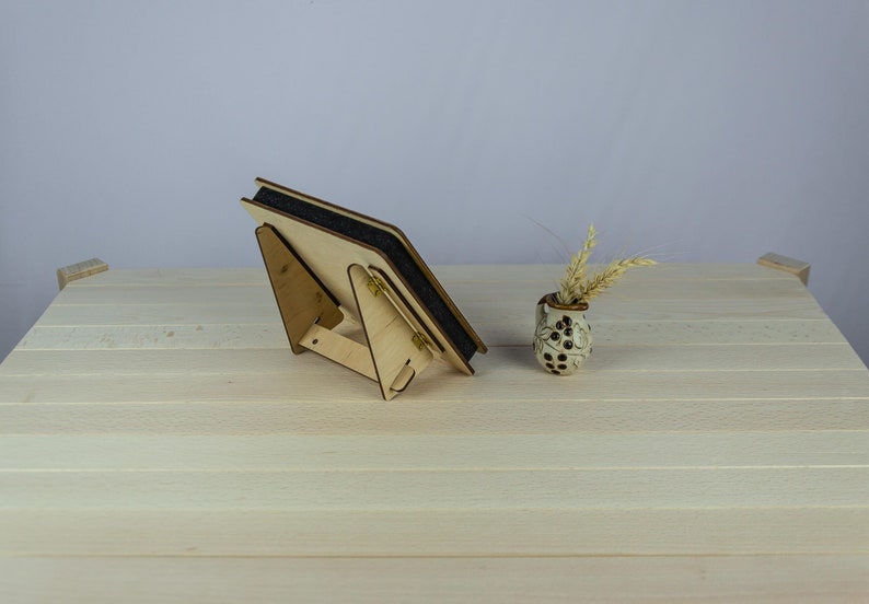 May include: A wooden picture frame with a black backing and a stand. The frame is on a light wood surface with a small vase with dried flowers.