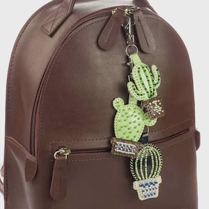 Puede incluir: Una mochila de cuero marrón con un llavero decorativo de cactus. El llavero presenta tres diseños de cactus verdes con detalles de cuentas y pequeñas macetas. La mochila tiene un asa superior y un bolsillo frontal con cremallera.