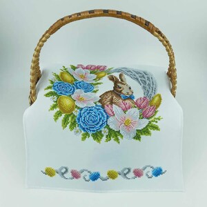 May include: A wicker basket with a curved handle, displaying a white cloth embroidered with an Easter design. The embroidery features a brown bunny, blue and pink flowers, yellow eggs, and a decorative border.