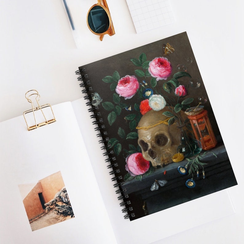 May include: Spiral-bound notebook featuring a still life painting with a skull, roses, and butterflies. The cover art has a dark background with a gold skull and pink and red roses. The notebook is open on a white surface.