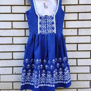 May include: A sleeveless blue dirndl dress featuring a white square neckline with white rickrack trim. The bodice and skirt hem are decorated with intricate white floral embroidery. A label inside reads Rose Dirndl with a small rose graphic.