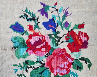 May include: A cross-stitch floral design featuring vibrant red, pink, and blue roses, with green leaves and accents. The embroidery is set against a neutral, textured fabric background, creating a rustic, handmade aesthetic.