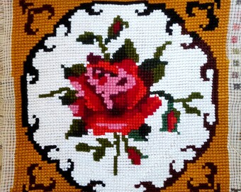 May include: A white needlepoint canvas with a red rose embroidered in the center. The rose is surrounded by green leaves and a brown border. The background is a solid yellow color.