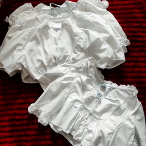 May include: A collection of white blouses featuring short puff sleeves, delicate lace trim along the necklines and cuffs, and button-down closures. These garments are crafted from a smooth, lightweight fabric and exhibit various neck styles.
