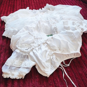 May include: A white blouse featuring puffed sleeves with sheer fabric panels and intricate lace trim details. The garment has a gathered neckline, a cinched bodice with a ruched front, and thin white drawstring ties hanging from the bottom hem.