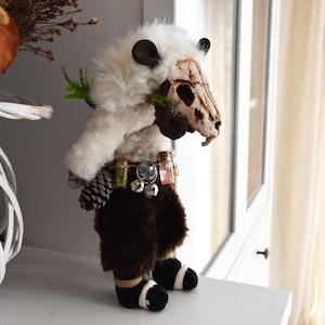 May include: A white and brown plush toy with a skull head, wearing a brown fur vest and a belt with small bottles and bells. The toy is holding a small branch in its hand.
