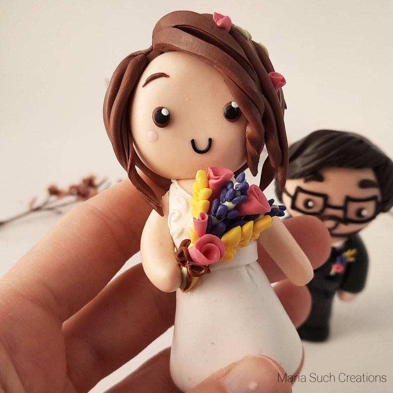 May include: A handmade polymer clay wedding cake topper featuring a bride and groom. The bride is wearing a white dress and holding a bouquet of yellow, purple, and pink flowers. The groom is wearing a black suit and a white shirt. The topper is a great way to add a personal touch to your wedding cake.