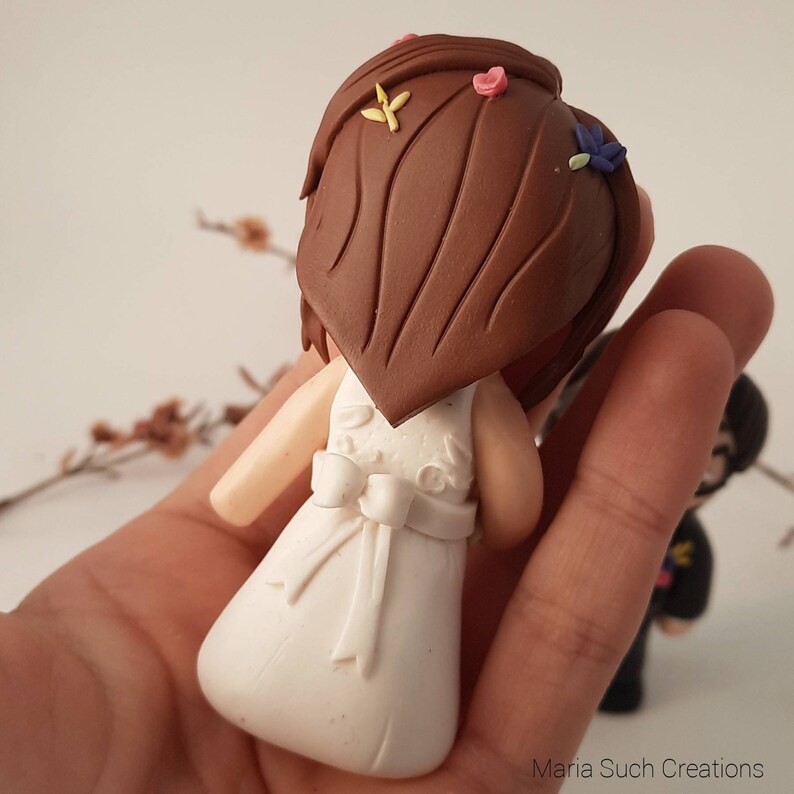 May include: A white wedding cake topper featuring a bride with brown hair and a white dress. The bride is wearing a white bow and has flowers in her hair.