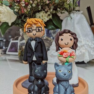 May include: A wedding cake topper featuring a bride and groom with black and gray cats. The groom is wearing a black suit and a white bow tie. The bride is wearing a white dress and holding a bouquet of flowers. The cats are sitting on a brown base.