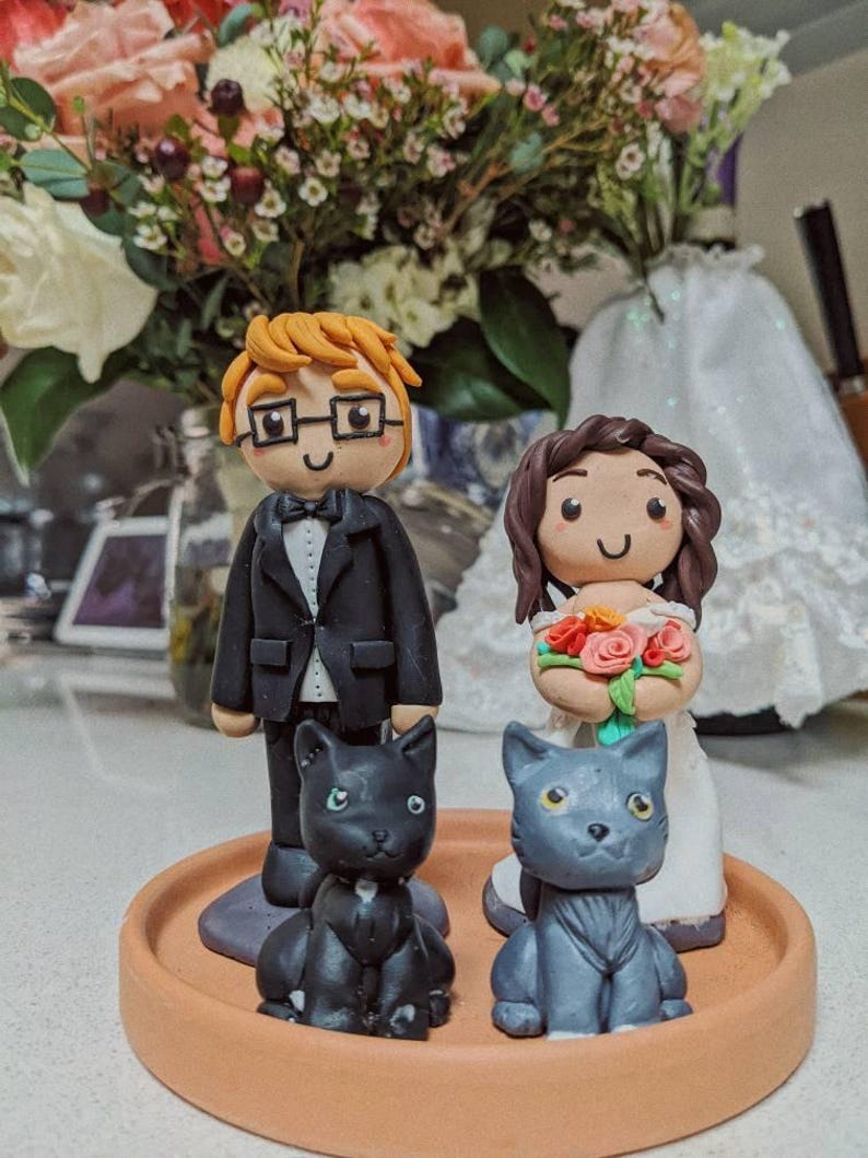 May include: A wedding cake topper featuring a bride and groom with black and gray cats. The groom is wearing a black suit and a white bow tie. The bride is wearing a white dress and holding a bouquet of flowers. The cats are sitting on a brown base.