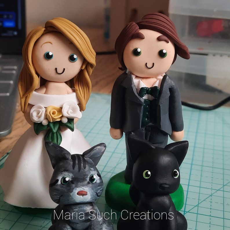 May include: A wedding cake topper set featuring a bride and groom figurine with a black cat figurine. The bride is wearing a white dress with a floral bouquet. The groom is wearing a black suit with a green vest and a bow tie. The cat is black with green eyes.