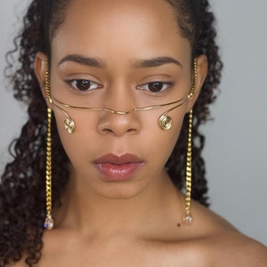 May include: A woman with curly dark brown hair is wearing a gold nose ring and matching long chain earrings with a crystal at the end. The nose ring is a thin wire with a spiral design on each side.