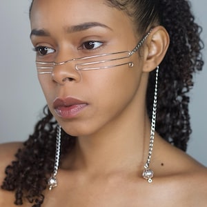 May include: Silver-toned face jewelry with a layered design across the nose and eyes, connected to chain earrings with decorative spheres. The jewelry is worn by a person with dark curly hair.