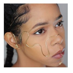 Puede incluir: Una mujer lleva una pieza de joyería de oro para la cara y la oreja. La joyería tiene forma de onda y se envuelve alrededor del lado de su cara, oreja y nariz.