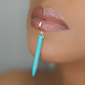 May include: A gold-colored lip ring with a turquoise-colored, elongated stone pendant. The jewelry is displayed on a person's lip, with a close-up view showcasing the details of the lip ring and the stone's texture.