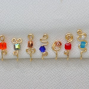 Op de afbeelding: Een verzameling van zeven goudkleurige neusmanchetten, elk met een uniek ontwerp en kleurrijke kraalaccenten. De kralen zijn in verschillende vormen en kleuren, waaronder oranje, iriserend, koper, blauw, rood en turquoise.