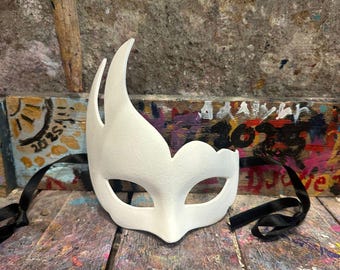 Può includere: Una maschera bianca con un design curvo unico. Presenta aperture per gli occhi e un naso a punta. Nastri neri sono attaccati per legarla. La maschera è posizionata su una superficie colorata e strutturata. Adatta per feste in maschera.