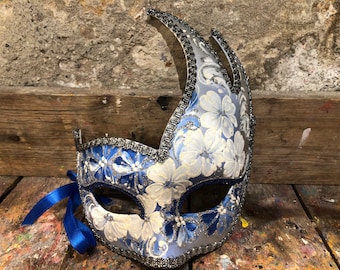 Può includere: Una maschera da carnevale blu e argento con motivi floreali bianchi. La maschera ha una parte superiore lunga, appuntita e curva e un nastro blu attaccato.