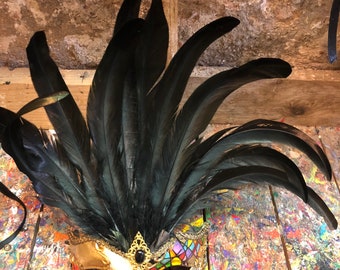 Può includere: Una maschera da carnevale con base dorata e bianca, decorata con motivi colorati che ricordano le vetrate. La maschera è sormontata da un grande pennacchio di piume nere.