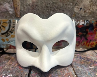 Può includere: Una maschera bianca e vuota per il ballo in maschera con aperture per gli occhi e un naso prominente. La maschera è realizzata in un materiale liscio e ha un design semplice. Un cordino nero è attaccato ai lati. La maschera è su una superficie di legno colorata e macchiata di vernice.