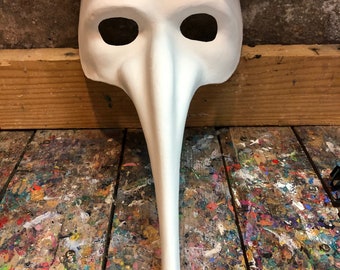 May include: White papier-mâché masquerade mask with a long, curved nose and eye holes. The mask is sitting on a wooden surface.
