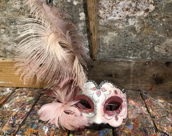 Può includere: Una maschera da carnevale rosa e argento con glitter e un delicato disegno floreale. La maschera è adornata con un grande boa di piume rosa.