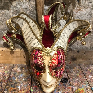 Könnte beinhalten: Eine rote und goldene venezianische Narrenmaske mit weißem Gesicht und Noten auf dem Hut. Die Maske hat ein schwarzes Band, das an der Rückseite befestigt ist.