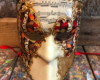 May include: A white and gold Venetian masquerade mask with red and brown accents. The mask features a musical score printed on the top half.