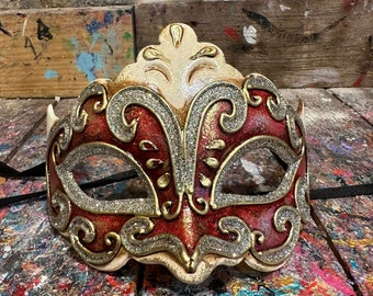 Può includere: Una maschera da carnevale rossa e dorata con dettagli intricati e accenti di glitter. La maschera ha uno stile veneziano tradizionale con una corona a punta e buchi per gli occhi.