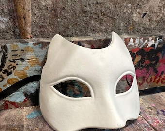 Può includere: Una maschera da ballo bianca sporca con orecchie da gatto e aperture per gli occhi. La maschera è realizzata in un materiale liscio ed è progettata per essere dipinta o decorata. Poggia su una superficie di legno usurata.