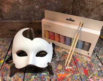 May include: A white masquerade mask with a diamond pattern on a wooden stand, next to a box of acrylic paint and two paintbrushes.