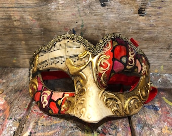 May include: An ornate Venetian mask with gold detailing, musical notes, and a red ribbon. The mask has a cream-coloured base with gold filigree and red, pink, and black stained glass-like details. The top edge is embellished with gold beads.