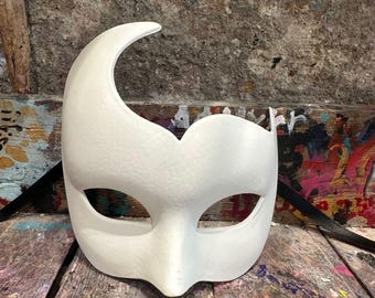 May include: A white, crescent-shaped masquerade mask with eye openings and a nose bridge. The mask is made of a smooth material and rests on a weathered, multi-colored wooden surface. The mask's design includes a curved top and a heart-shaped detail.