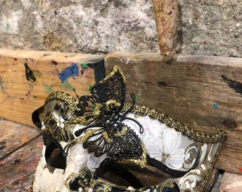 May include: A white and black masquerade mask with gold glitter trim and a black butterfly design. The mask has a floral pattern and is decorated with small white beads.