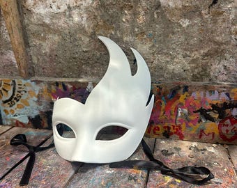 Può includere: Una maschera bianca da ballo in maschera con un design a forma di fiamma sulla parte superiore sinistra. La maschera ha aperture per gli occhi ed è legata con nastri neri. Poggia su una superficie di legno con schizzi di vernice colorati.