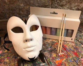 May include: A white, blank, masquerade mask with a diamond pattern and black trim. The mask is on a stand. A set of paint tubes and two paint brushes are in the background.