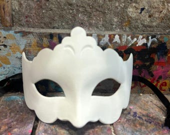 Può includere: Una maschera bianca decorativa con bordo smerlato e aperture per gli occhi. La maschera ha un elemento decorativo centrale ed è attaccata a un cinturino nero. La maschera è probabilmente per un evento in costume.