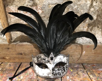 Può includere: Una maschera da ballo in maschera decorativa con un design floreale bianco e argento, accentuata da piume nere. La maschera presenta una gemma nera e un nastro nero, perfetta per una festa in costume o un ballo in maschera.