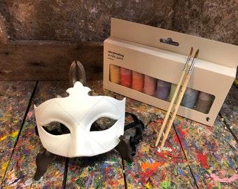 May include: A white masquerade mask with a diamond pattern on a wooden surface with paint splatters.  A box of acrylic paint with 8 tubes of paint and two paintbrushes are also on the surface.