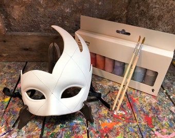 May include: A white plastic masquerade mask with a diamond pattern and eye holes. The mask is on a stand and there is a box of paint and brushes in the background.
