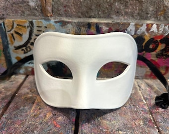 Può includere: Una semplice maschera bianca per ballo in maschera con fori per gli occhi e un ponte nasale. Sono attaccati nastri neri per fissarla. La maschera è appoggiata su una superficie di legno multicolore.