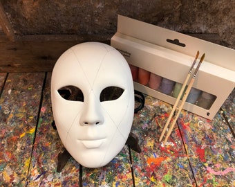 Può includere: Una maschera bianca vuota con linee a forma di diamante disegnate su di essa. La maschera è su una superficie di legno con schizzi di vernice. Una scatola di vernice acrilica e due pennelli sono sulla superficie.
