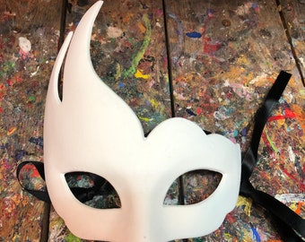 May include: A white masquerade mask with a curved design and eye openings. The mask has black ribbon ties and is set against a colorful, paint-splattered wooden surface. The mask is designed for a costume or party.
