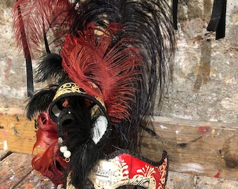 May include: A masquerade mask with black and red feathers, a white and red design, and black trim. The mask has a musical note pattern and is decorated with gold accents.