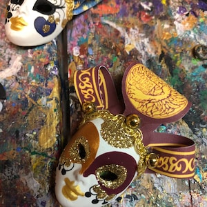 Peut inclure: Deux masques de carnaval vénitiens décoratifs avec des paillettes dorées et des détails colorés. Les masques sont en céramique et ont une base blanche avec des accents dorés et bordeaux. Un masque a un motif bleu et or avec des étoiles, tandis que l'autre a un motif bordeaux et or avec un ruban doré.