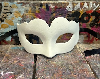 Può includere: Una maschera bianca da ballo in maschera con bordi smerlati, con aperture per gli occhi e un naso a punta. La maschera è realizzata in un materiale liscio e ha un nastro nero attaccato. Poggia su una superficie di legno colorata e macchiata di vernice.